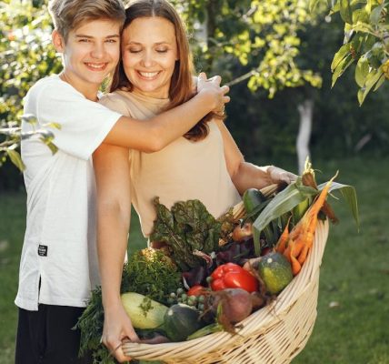 Mother and son smiling outdoors with a basket full of fresh vegetables, embracing in a sunny garden.