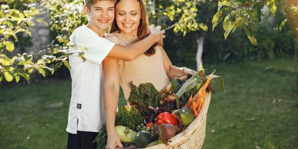 Mother and son smiling outdoors with a basket full of fresh vegetables, embracing in a sunny garden.